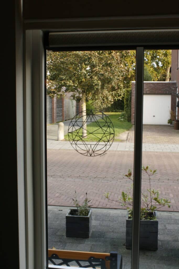 Foto gemaakt vanuit binnenshuis. Kozijn zichtbaar met op het raam basis krachtsymbool Ebonaq. Plantenbakken, straat, bomen en een garage