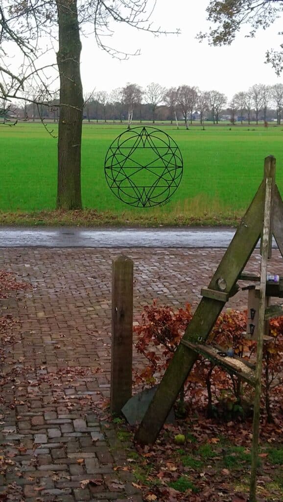 Een foto waar het uitzicht naar buiten wordt getoond. Een inrit, met een straat en daarachter een boom en weiland. Op het raam zit het krachtsymbool Ebonaq opgeplakt.